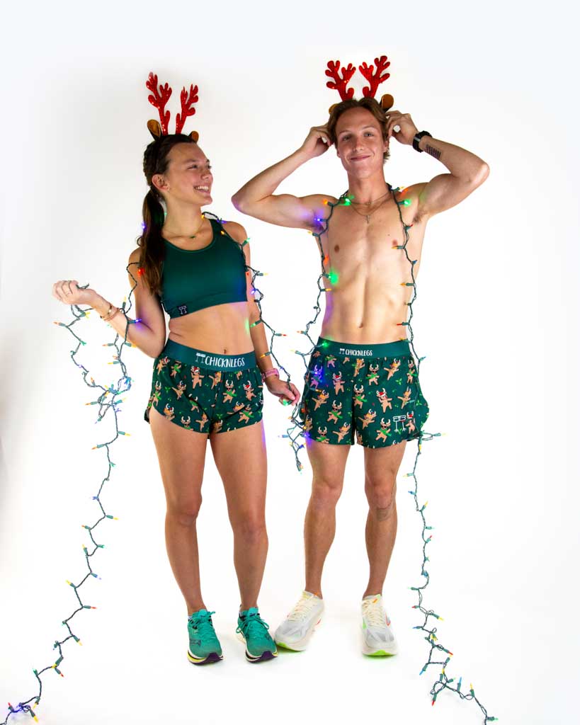 Full body view of model wearing ChicknLegs Women’s 1.5-inch split running shorts featuring a soft elastic waistband, rear zipper pocket, and black mesh liner for full coverage and breathability and Men's 4 inch split running shorts, both featured are reindeer dancing with santa hats and christmas lights, paired with a forest green sports bra.