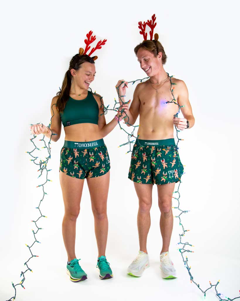 Full body view of model wearing ChicknLegs Women’s 1.5-inch split running shorts featuring a soft elastic waistband, rear zipper pocket, and black mesh liner for full coverage and breathability and Men's 4 inch split running shorts, both featured are reindeer dancing with santa hats and christmas lights, paired with a forest green sports bra.