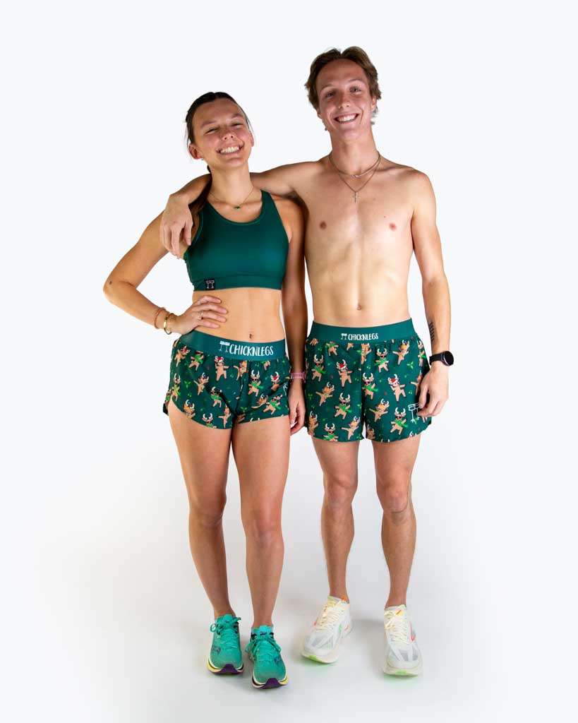 Full body view of model wearing ChicknLegs Women’s 1.5-inch split running shorts featuring a soft elastic waistband, rear zipper pocket, and black mesh liner for full coverage and breathability and Men's 4 inch split running shorts, both featured are reindeer dancing with santa hats and christmas lights, paired with a forest green sports bra.
