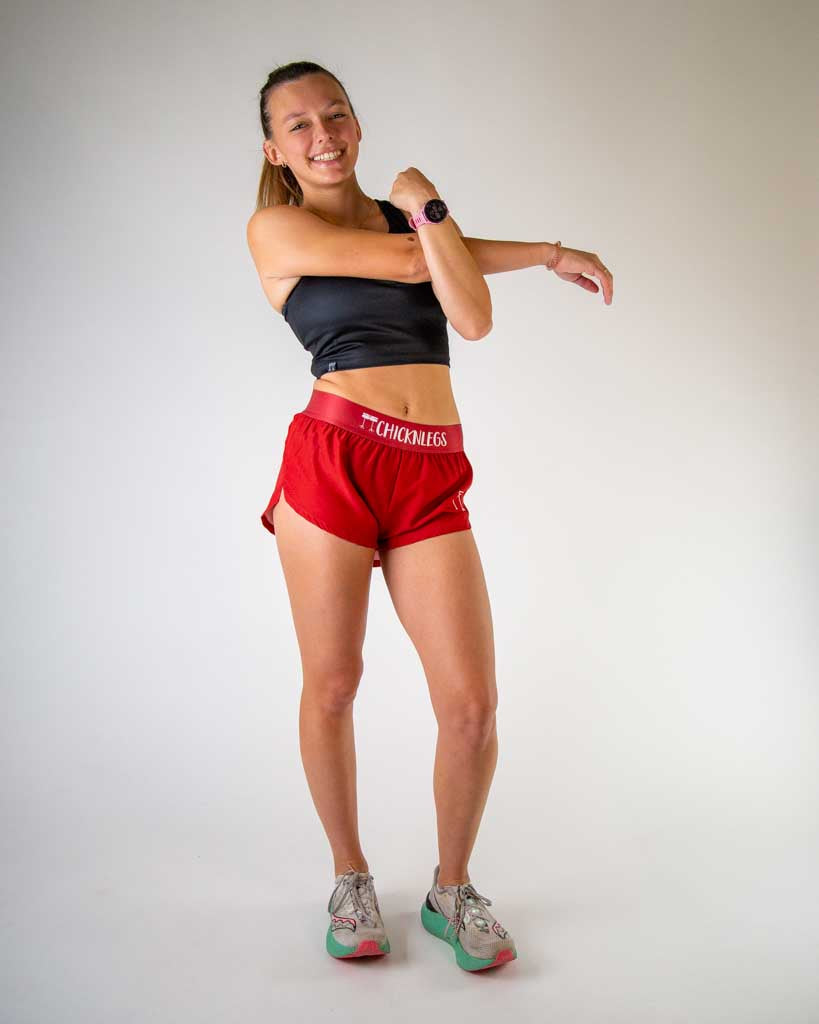 Full body view of model wearing ChicknLegs women’s 1.5-inch split running shorts in solid cardinal red, styled with black crop top and white running shoes.