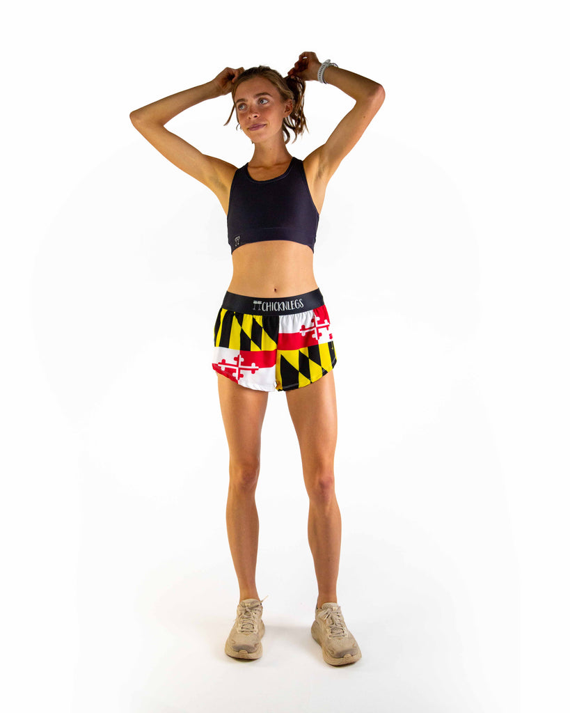 Full body view of model wearing ChicknLegs Women’s 1.5-inch split running shorts featuring a soft elastic waistband, rear zipper pocket, and black mesh liner for full coverage and breathability. Featured is the Maryland State flag paired with a black sports bra.