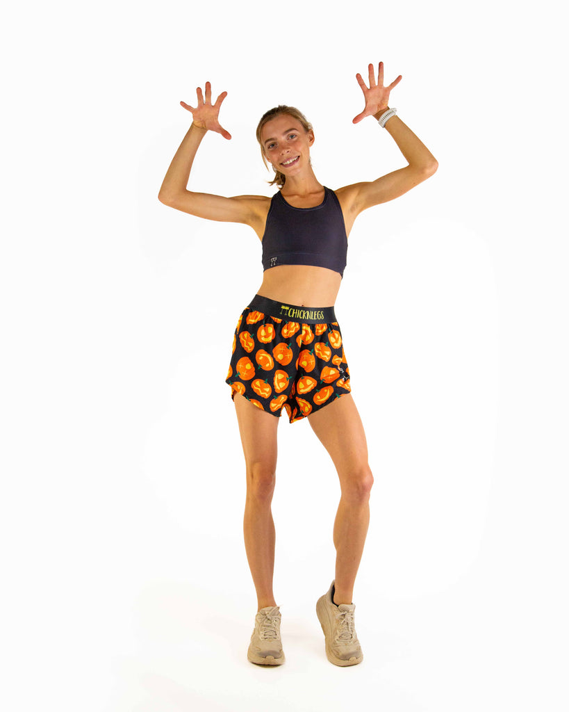 Full body view of model wearing ChicknLegs Women’s 4-inch split running shorts with soft elastic waistband, featured in the Pumpkins design, with jack o lantern pattern and black waistband with yellow text to match.