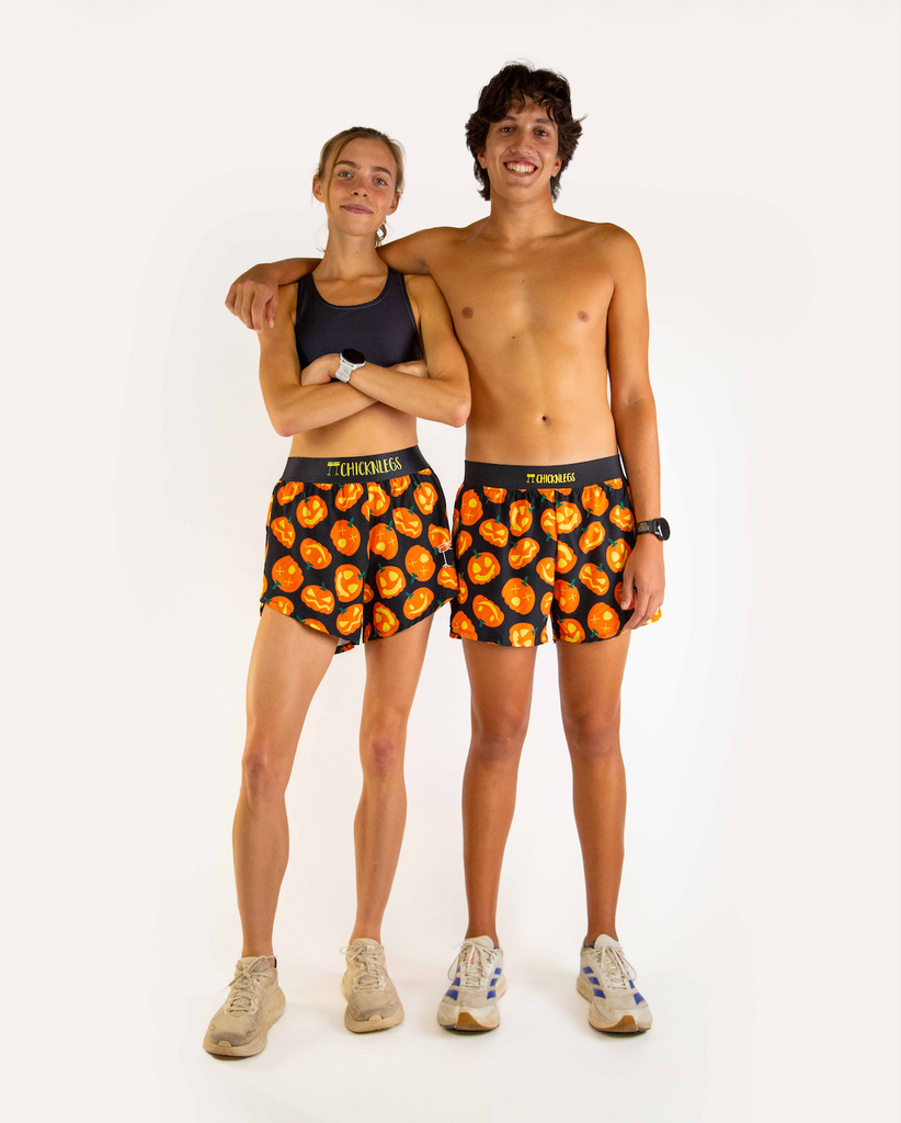 Both models are wearing ChicknLegs in the pumpkin or jack o lantern design, matching in the men's 4 inch split runnning shorts and women's 4 inch shorts. Product is styled with black sports bra and white shoes.