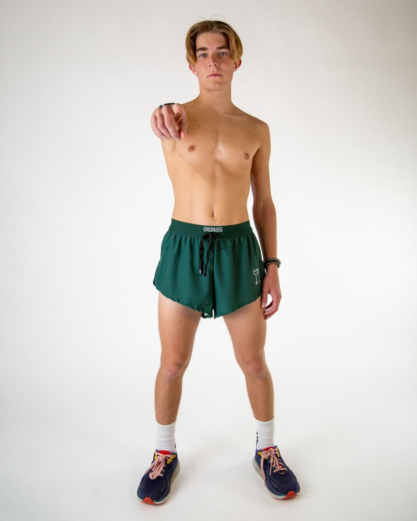 Full body view of model wearing ChicknLegs men’s 2-inch split running shorts in solid forest green, styled with black running shoes.