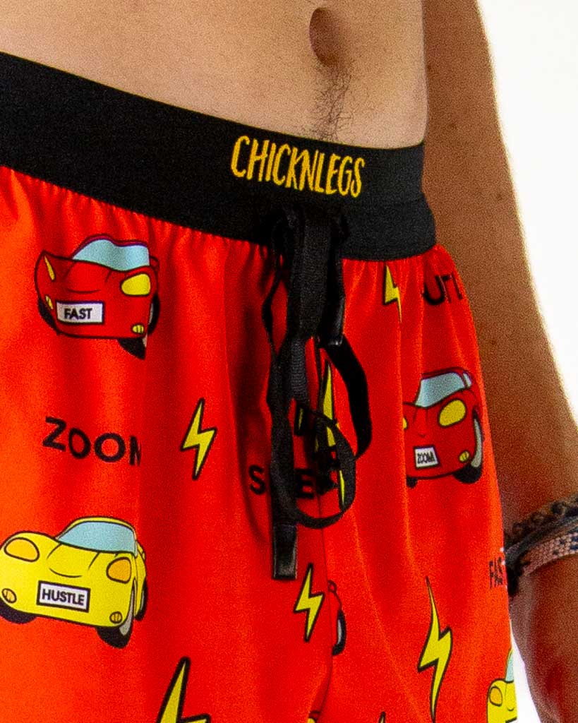 Close-up detail of ChicknLegs men’s 2-inch running shorts, orange and red fabric with yellow lightning bolt pattern, red and yellow cars and drawstring waistband.