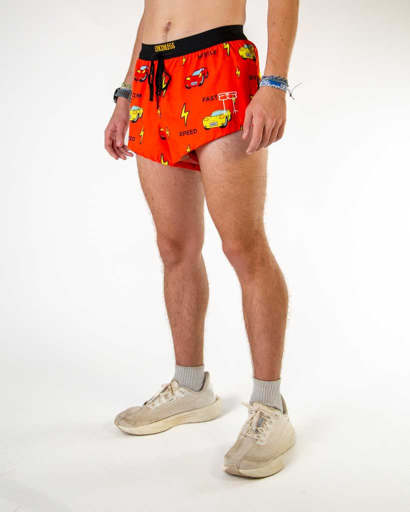 Left side view of ChicknLegs men’s 2-inch split running shorts in red orange with yellow lightning bolts, red and yellow cars that say SPEED, HUSTLE, and FAST featuring split leg design for running.