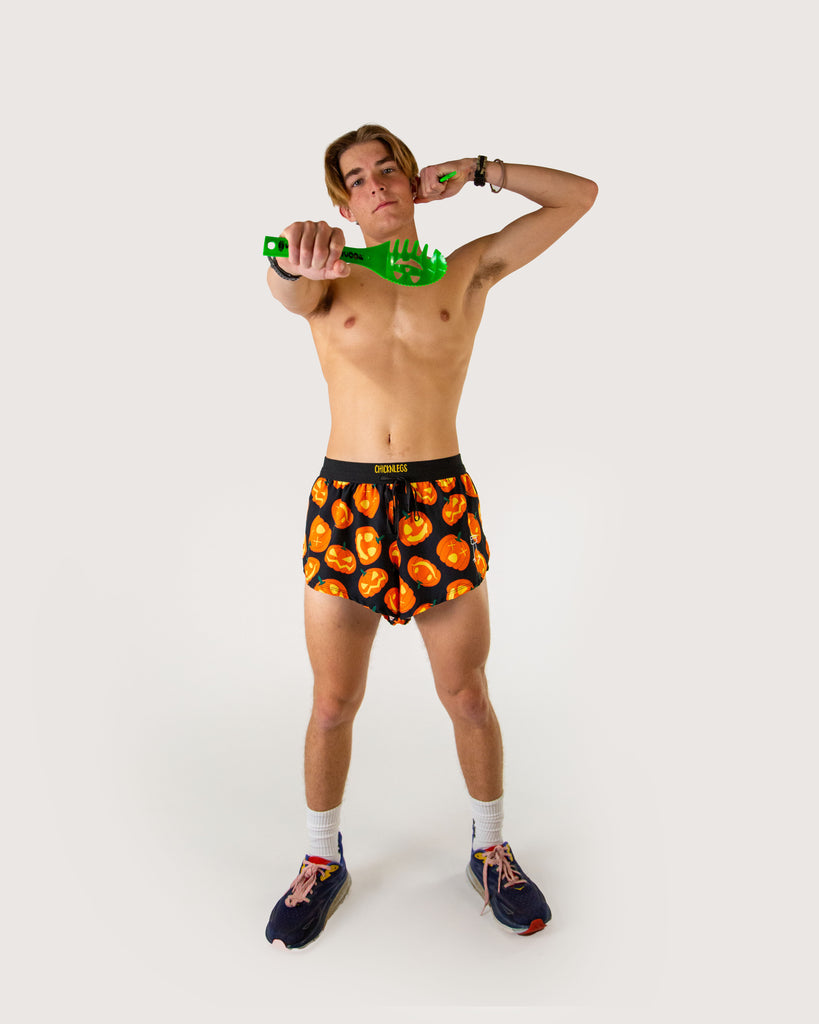 Full body view of model wearing ChicknLegs men’s 2-inch split running shorts in black background, orange pumpkins, styled with black running shoes and holding carving tools for pumpkins.