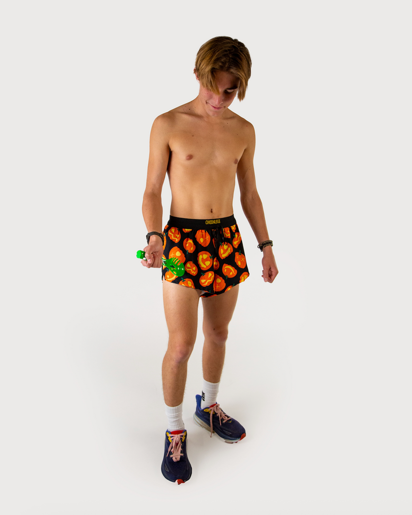 Full body view of model wearing ChicknLegs men’s 2-inch split running shorts in black background, orange pumpkins, styled with black running shoes.