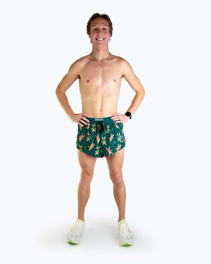 Full body view of model wearing ChicknLegs Men’s 2-inch DS Split Running Shorts featuring a soft elastic drawstring waistband, inner pocket, and black mesh liner for full coverage and breathability. Featured are the rockin reindeer design in christmas lights and santa hats with a dark green background.