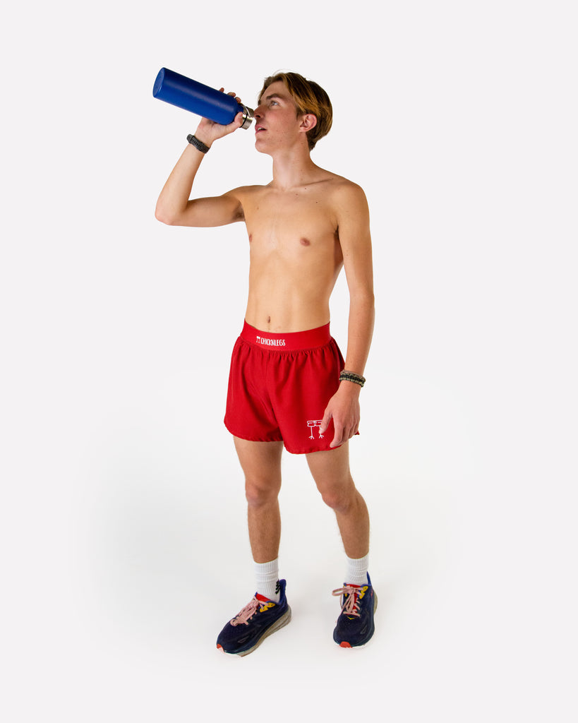 Full body view of model wearing ChicknLegs men’s 4-inch split running shorts in solid cardinal red, styled with black running shoes.