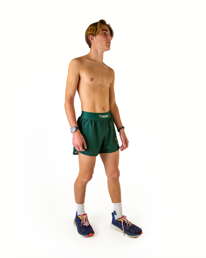 Full body view of model wearing ChicknLegs men’s 4-inch split running shorts in solid forest green, styled with black running shoes.