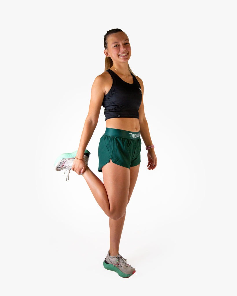 Full body view of model wearing ChicknLegs women’s 1.5-inch split running shorts in solid forest green, styled with black crop top and white running shoes.