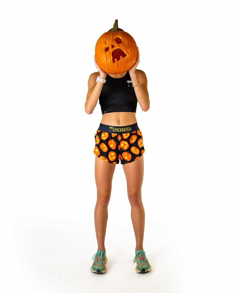 Full body view of model wearing ChicknLegs women’s 1.5-inch split running shorts with black background and Jack-o’-lantern pumpkin design, black waistband with yellow logo text, styled with running shoes.