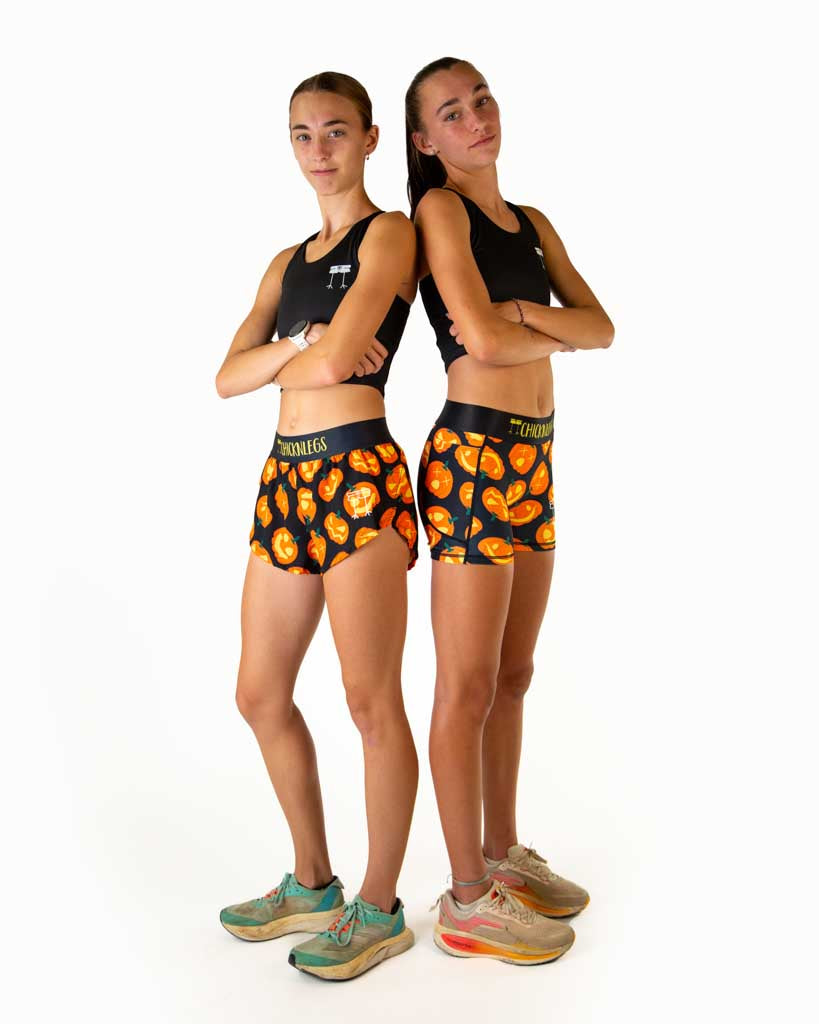 Full body view of model wearing ChicknLegs women’s 3-inch compression shorts in black with Jack-o’-lantern pumpkin design, black waistband with yellow text, styled with running shoes while paired with another runner wearing women's 1.5 inch split running shorts in the pumpkin design.