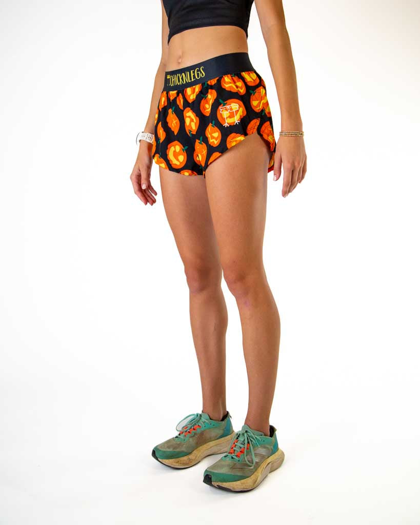 Left side view of ChicknLegs women’s 1.5-inch split running shorts in black with carved pumpkin Jack-o’-lantern design, featuring split leg cut and black waistband with yellow text.