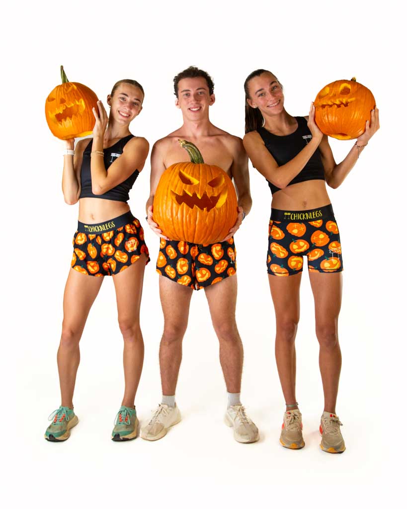 Full body view of model swearing ChicknLegs women’s 3-inch compression shorts, women's 1.5 inch split running shorts and the men's 2 inch drawstring shorts in black with Jack-o’-lantern pumpkin design, black waistband with yellow text, styled with running shoes.
