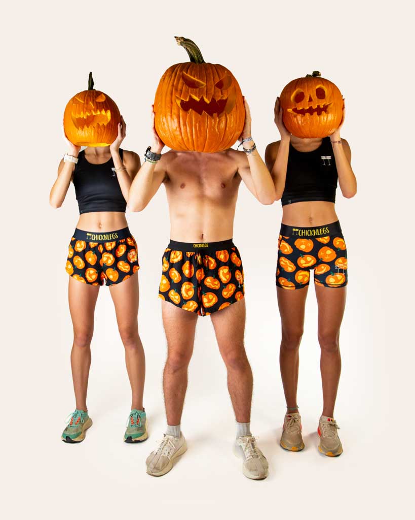 Full body view of models wearing ChicknLegs women’s 1.5-inch split running shorts and men's 2 inch drawstring split running shorts  and women's 3 inch compression shorts with black background and Jack-o’-lantern pumpkin design, black waistband with yellow logo text, styled with running shoes.