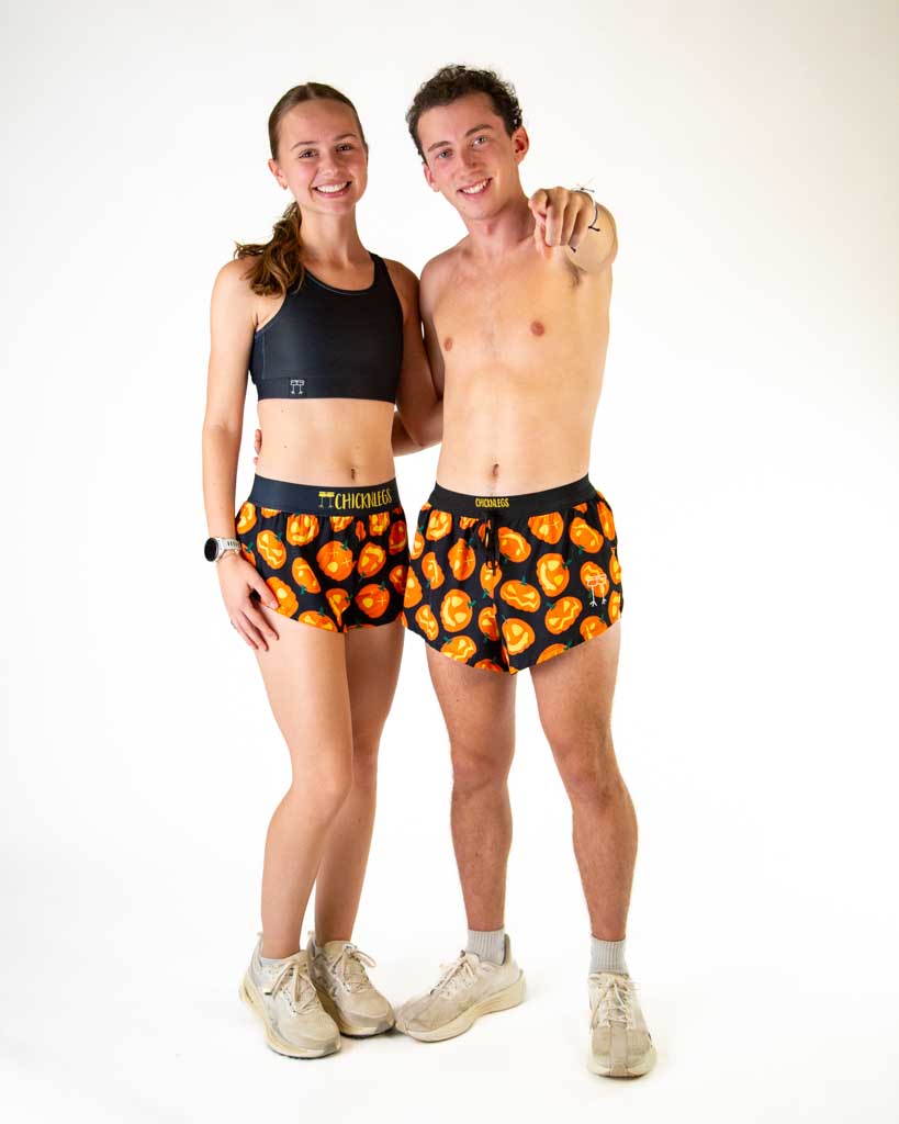 Full body view of models wearing ChicknLegs women’s 1.5-inch split running shorts and men's 2 inch drawstring split running shorts with black background and Jack-o’-lantern pumpkin design, black waistband with yellow logo text, styled with running shoes.