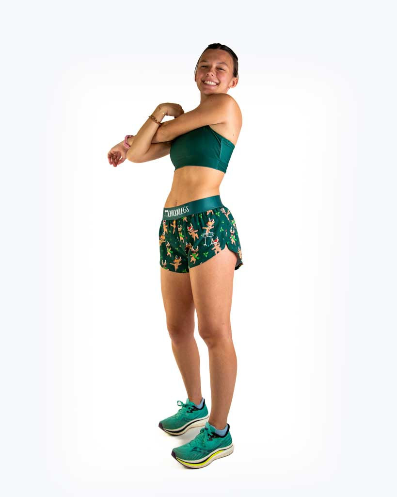 Full body view of model wearing ChicknLegs Women’s 1.5-inch split running shorts featuring a soft elastic waistband, rear zipper pocket, and black mesh liner for full coverage and breathability. Featured are reindeer dancing with santa hats and christmas lights, paired with a forest green sports bra.
