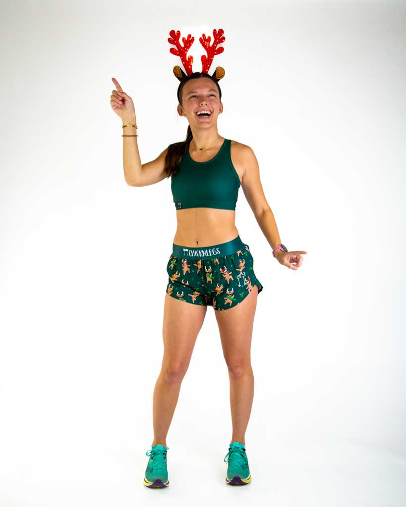 Full body view of model wearing ChicknLegs Women’s 1.5-inch split running shorts featuring a soft elastic waistband, rear zipper pocket, and black mesh liner for full coverage and breathability. Featured are reindeer dancing with santa hats and christmas lights, paired with a forest green sports bra and reindeer ears.