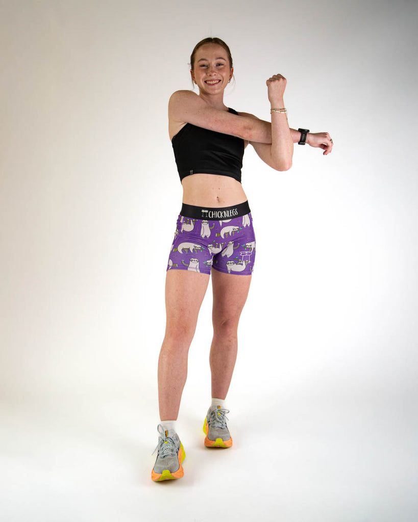 Full body view of ChicknLegs women’s 3-inch compression spandex running shorts with a purple background and sloths all over, with side pockets. Paired with a black crop top.