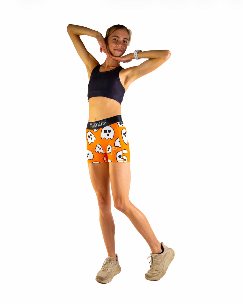 Full body view of model wearing ChicknLegs Women’s 3-inch compression shorts featuring a soft elastic waistband, dual side pockets, and spandex-polyester fabric for a supportive, chafe-free fit. Paired with a black sports bra to match the Ghosts with an orange background and black waistband.