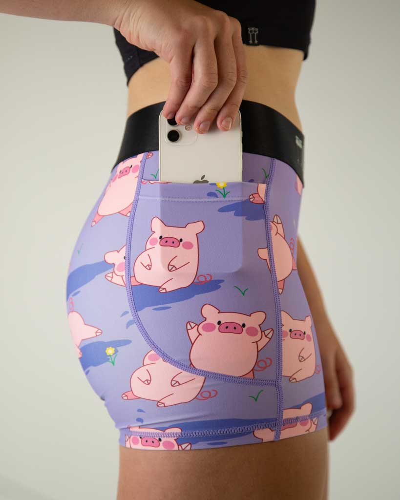 A close shot of ChicknLegs women’s 3-inch compression spandex with side pockets, lilac purple background and pink piggies design with phone in pocket.