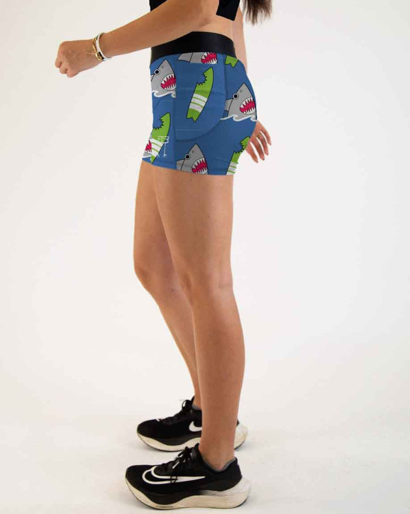 Runner is wearing ChicknLegs in the women's 3 in compression spandex with pockets shorts in the blue sharks design. The print has sharks and green surfboards on it with an ocean blue background.