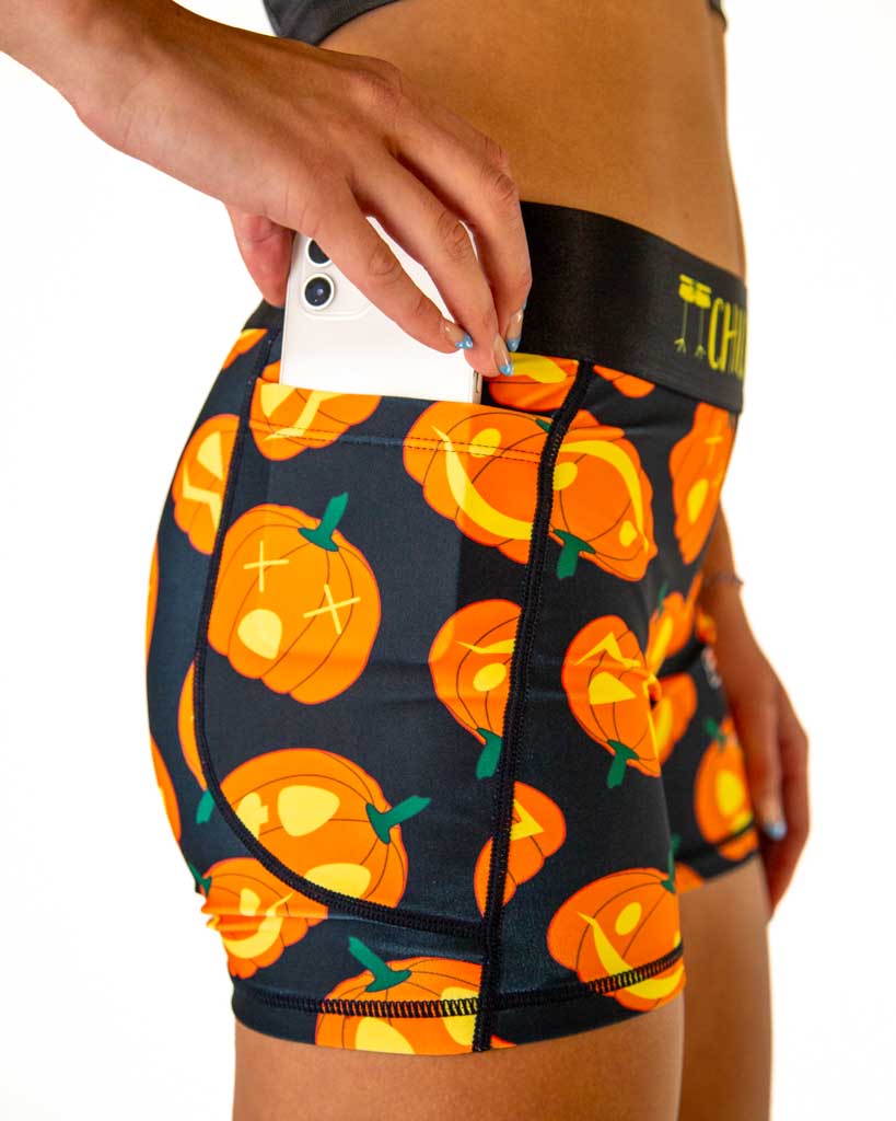 Close-up of ChicknLegs women’s 3-inch compression shorts, black fabric with orange Jack-o’-lantern pumpkin design, black waistband with yellow text, and side pocket.