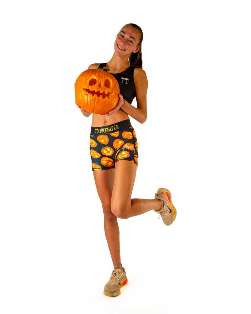 Full body view of model wearing ChicknLegs women’s 3-inch compression shorts in black with Jack-o’-lantern pumpkin design, black waistband with yellow text, styled with running shoes and holding a carved pumpkin.