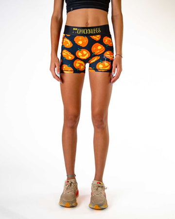 Front view of ChicknLegs women’s 3-inch compression shorts with side pockets, black background with Jack-o’-lantern pumpkin design, black waistband with yellow text.