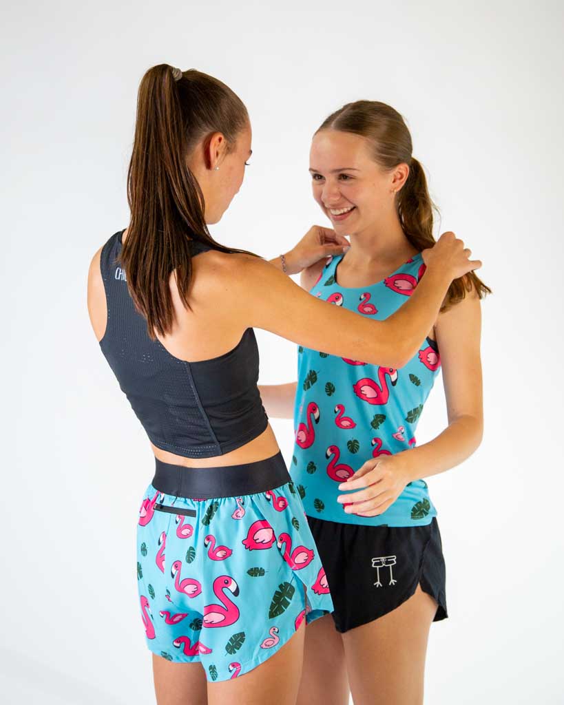 A full body image is shown of the runners wearing ChicknLegs black crop top and women's 4 inch split running shorts in the Blue Flamingo design, and the Blue Flamingo performance running singlet, matching with bright neon shoes.