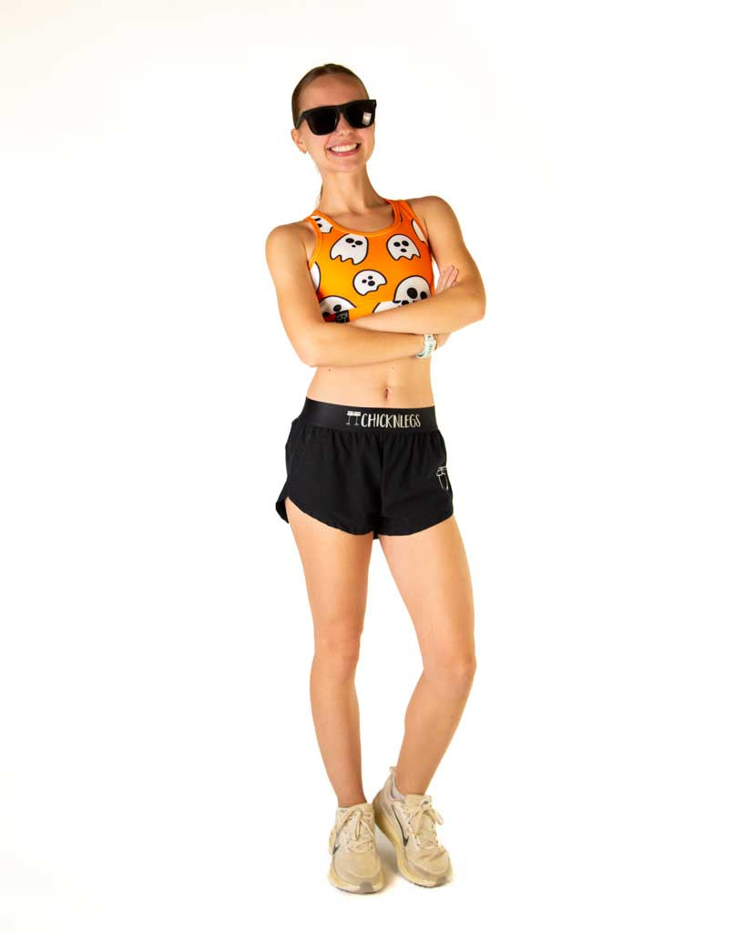 Full body view of model wearing ChicknLegs women’s OG sports bra with an orange background and white ghosts, styled with white shoes and black sunglasses.