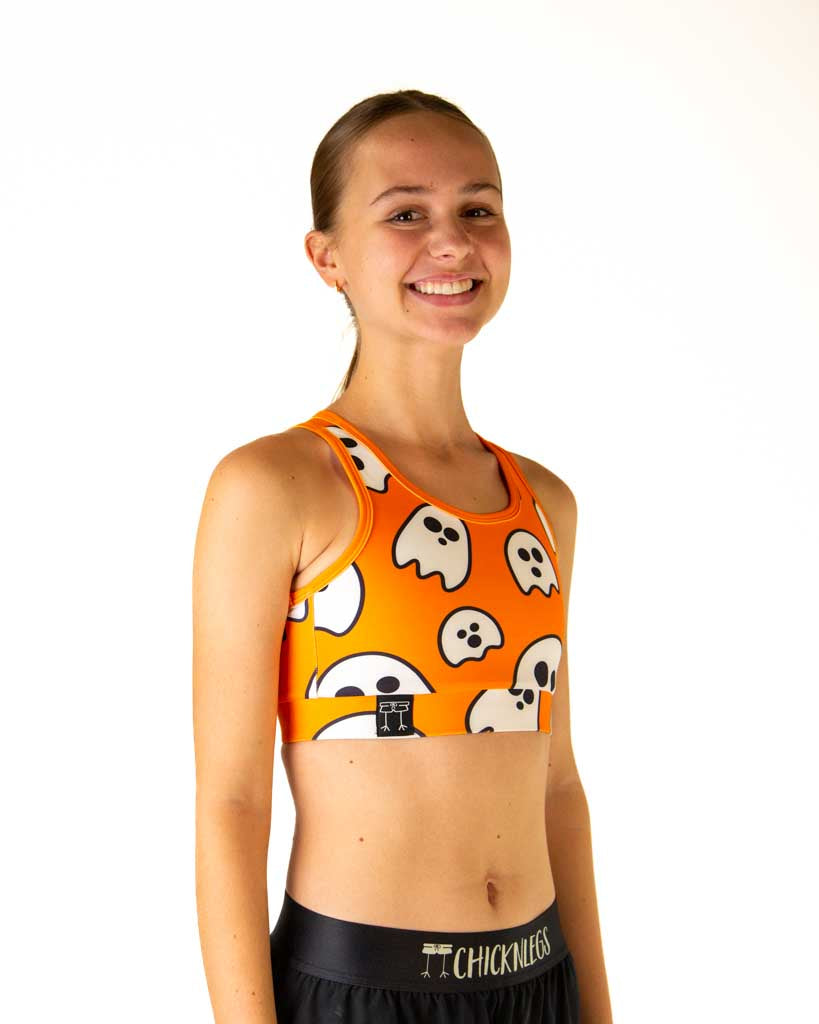 Right side view of ChicknLegs women’s OG sports bra in orange with white ghosts, highlighting logo on underband of the bra.