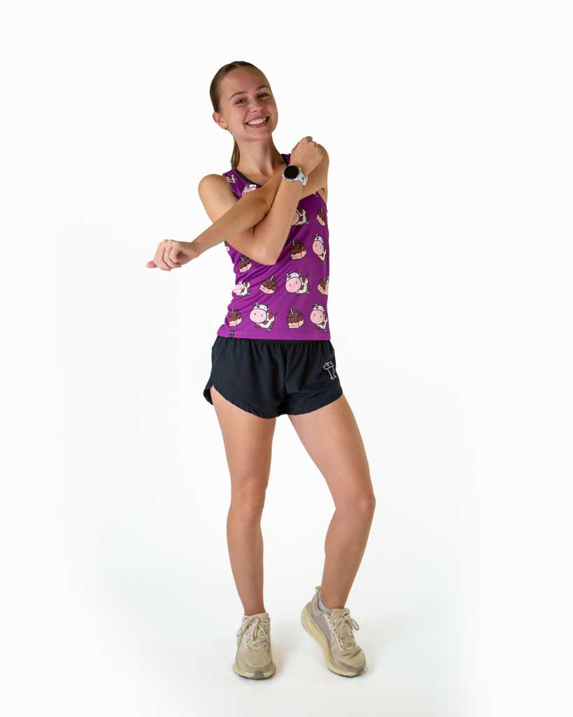 Full body view of runner wearing ChicknLegs in the women's performance running singlet in the choccy cow design and purple background, while stretching and wearing white runners shoes.