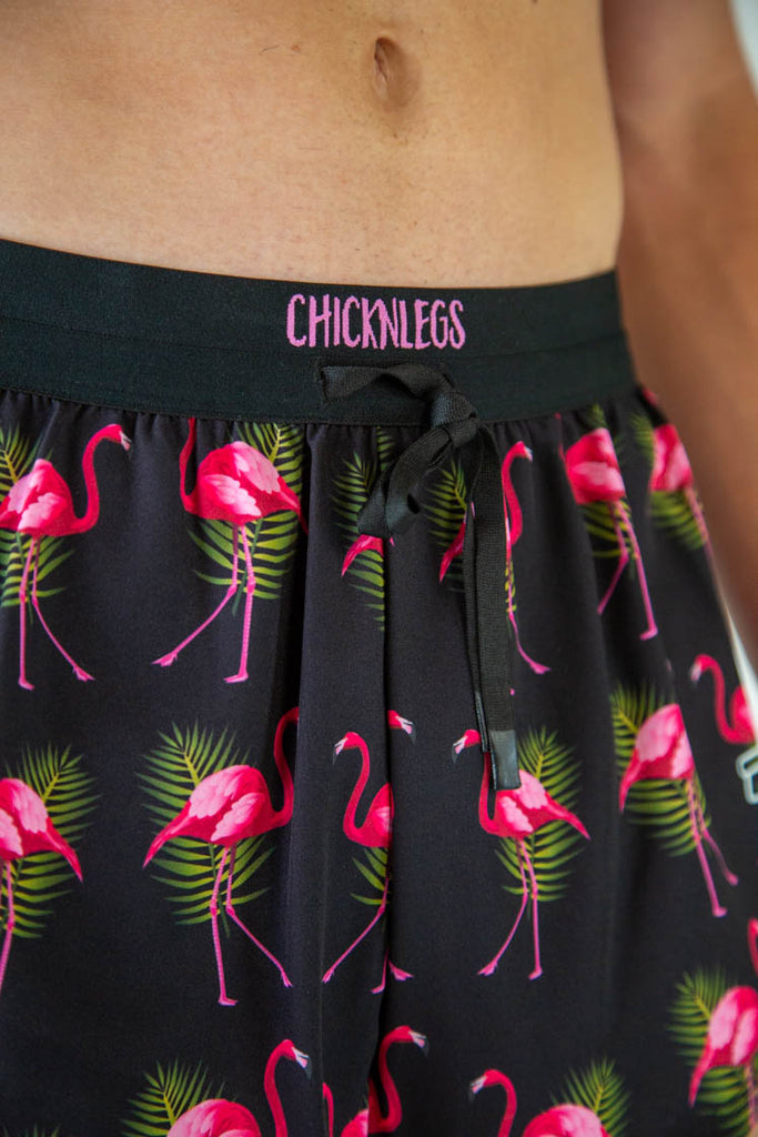 ChicknLegs Men's 2" DS drawstring shorts are shown on the runner. The print has hot pink flamingos with green leaves and a black background on it. A close up show is shown of the drawstrings and front logo.