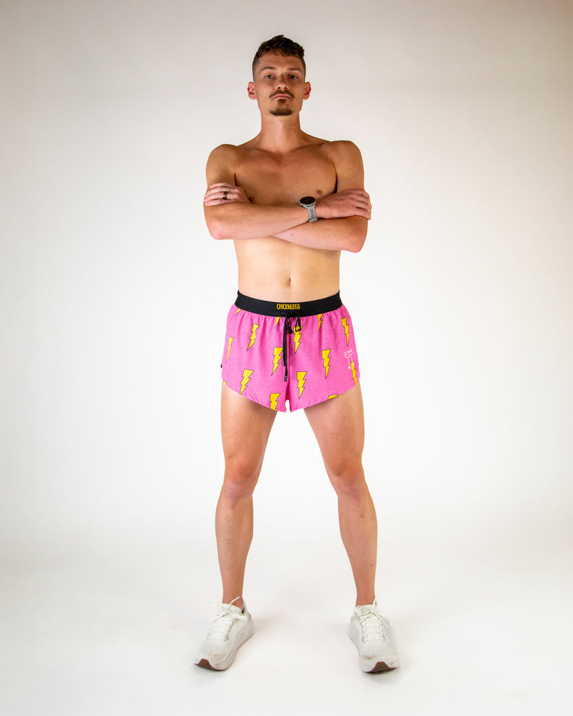 Full body view of model wearing ChicknLegs men’s 2-inch split running shorts with drawstring in hot pink with yellow lightning bolts, styled with white running shoes.