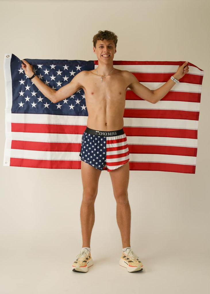 Model wearing Chicknlegs in men's 2 inch split running shorts in the usa design, facing front while posing with the USA flag.