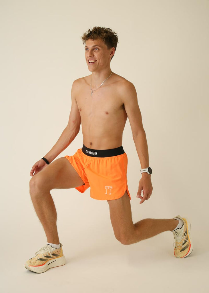 Model wearing Chicknlegs men's 4 inch split running shorts in the neon orange design, posing while stretching.