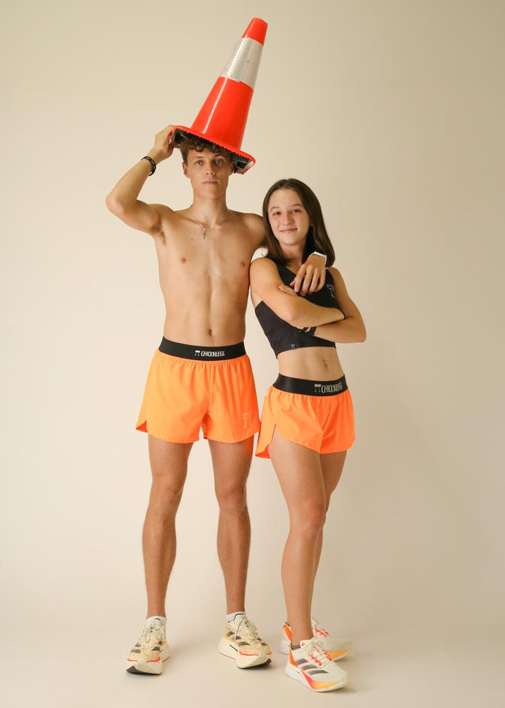 Model wearing Chicknlegs men's 4 inch split running shorts in the neon orange design and model wearing women's 1.5 inch chicknlegs split running shorts in neon orange design, posing together with bright orange cone.