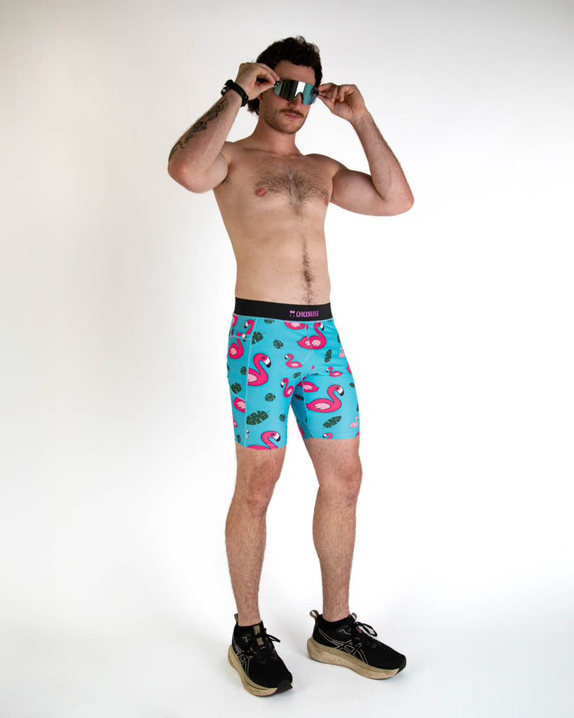 Full body view of ChicknLegs men's 8-inch compression spandex running half tights with breathable liner, blue background and pink flamingos, with a black waistband and side pockets. Paired with black shoes and blue sunglasses.