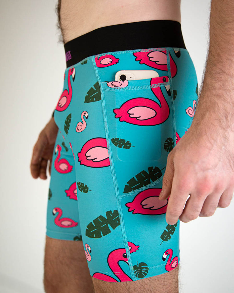 A close up view of ChicknLegs men's 8-inch compression spandex running half tights with breathable liner, blue background and pink flamingos, with a black waistband. A phone is shown inside the pocket.