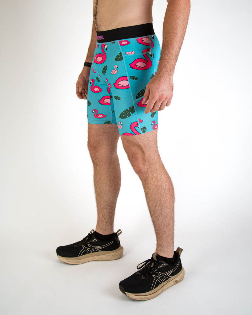 Left side view of ChicknLegs men's 8-inch compression spandex running half tights with breathable liner, blue background and pink flamingos, with a black waistband and side pockets. Paired with black shoes.