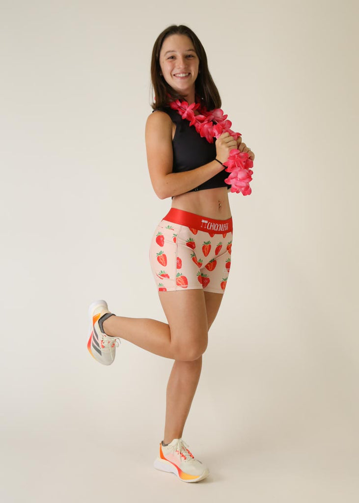 Model is wearing Chicknlegs women's 3 inch compression shorts for running in the strawberry szn design in full body shot posing with pink flowers.
