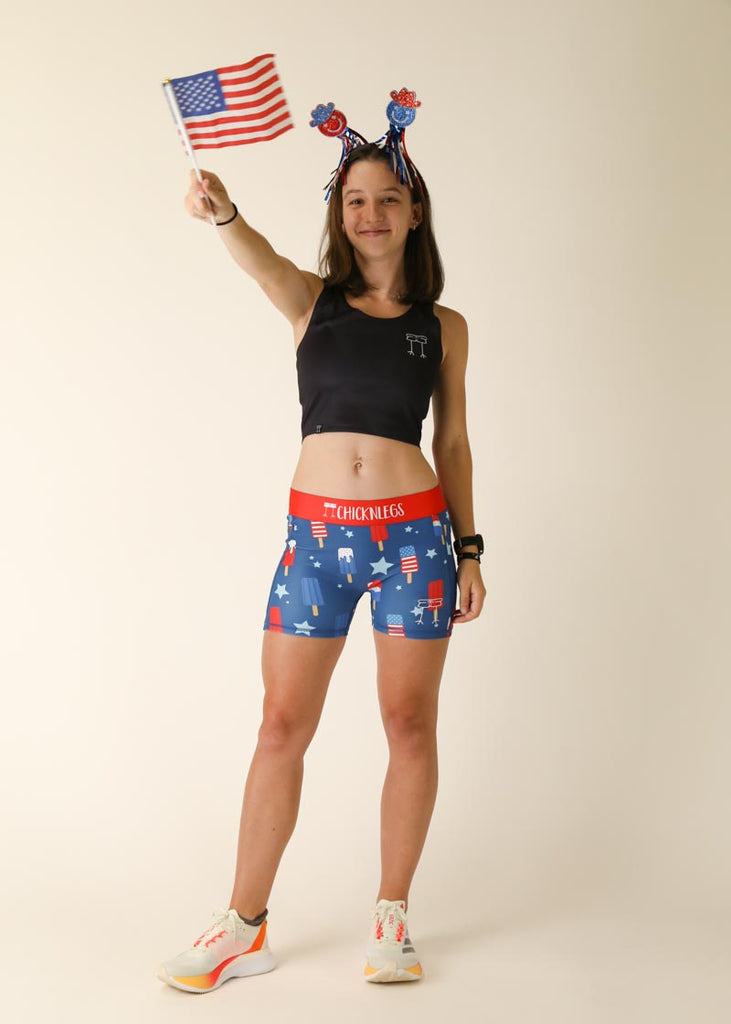 Model is wearing Chicknlegs women's 3 inch compression shorts for running in the usa popsicles design, posing with a USA flag.