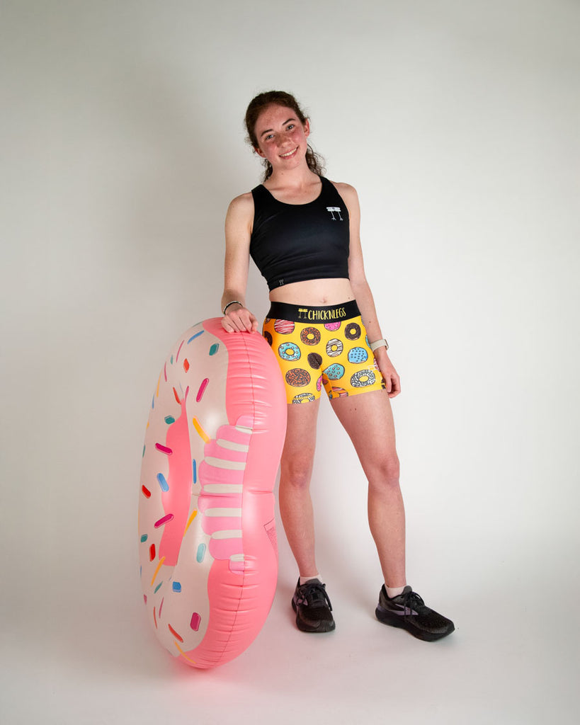 Runner is wearing ChicknLegs in the women's 3 in compression spandex shorts in the salty donuts design. Fun print with food in blue, brown, pink, yellow, red, orange, green, colors! Model is holding a giant donut with sprinkles on it.