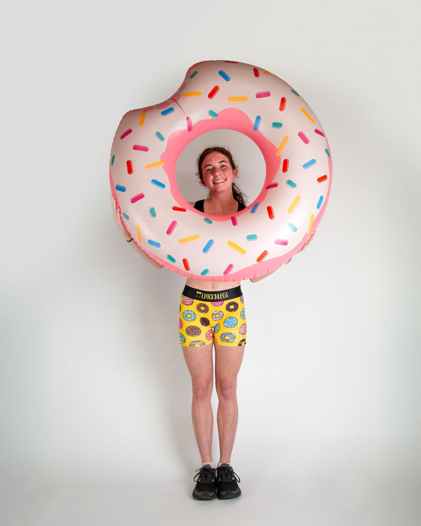 Runner is wearing ChicknLegs in the women's 3 in compression spandex shorts in the salty donuts design. Fun print with food in blue, brown, pink, yellow, red, orange, green, colors! Model is holding a giant donut with sprinkles on it.