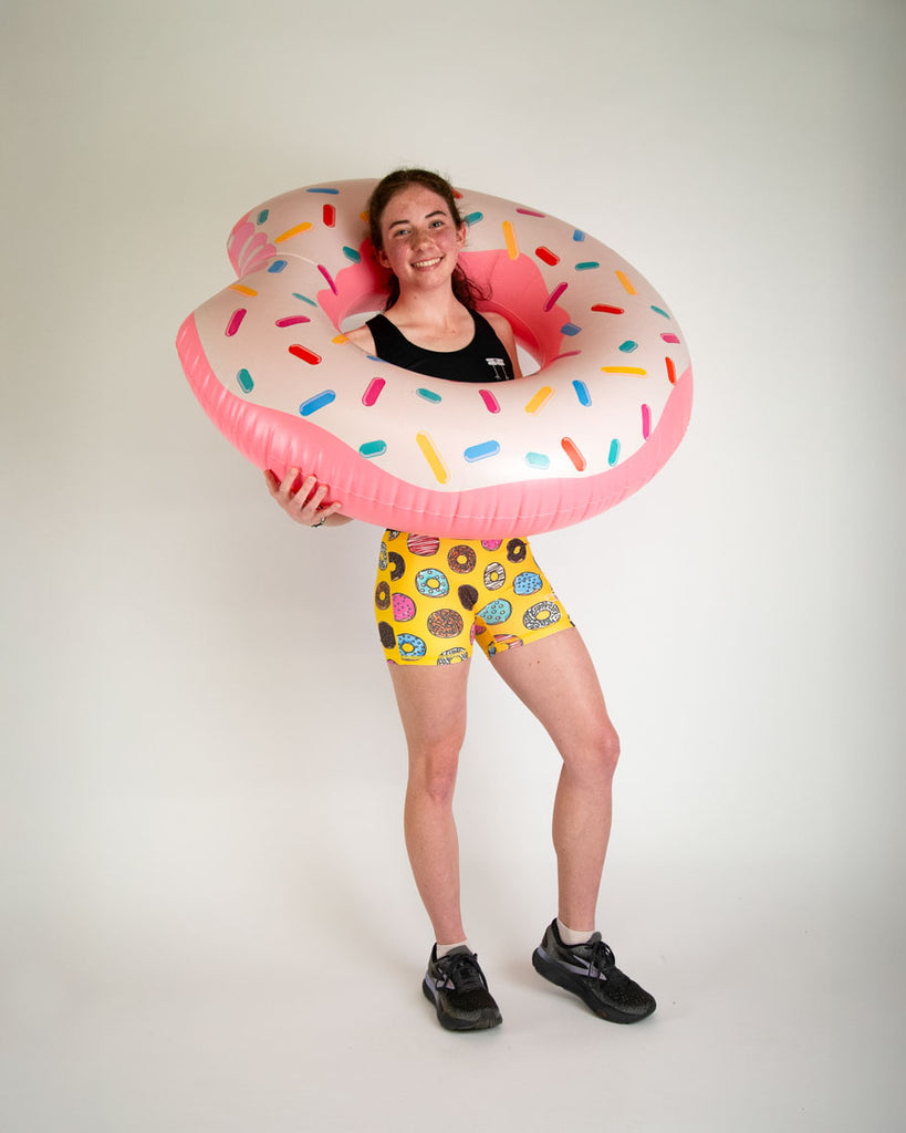 Runner is wearing ChicknLegs in the women's 3 in compression spandex shorts in the salty donuts design. Fun print with food in blue, brown, pink, yellow, red, orange, green, colors! Model is holding a giant donut with sprinkles on it.