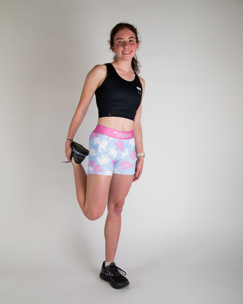 Runner is wearing ChicknLegs in the women's 3 in compression and spandex in the animal cookies design. The print has pink, white and blue on it with fun rainbow sprinkles on it. The model is stretching while posing.
