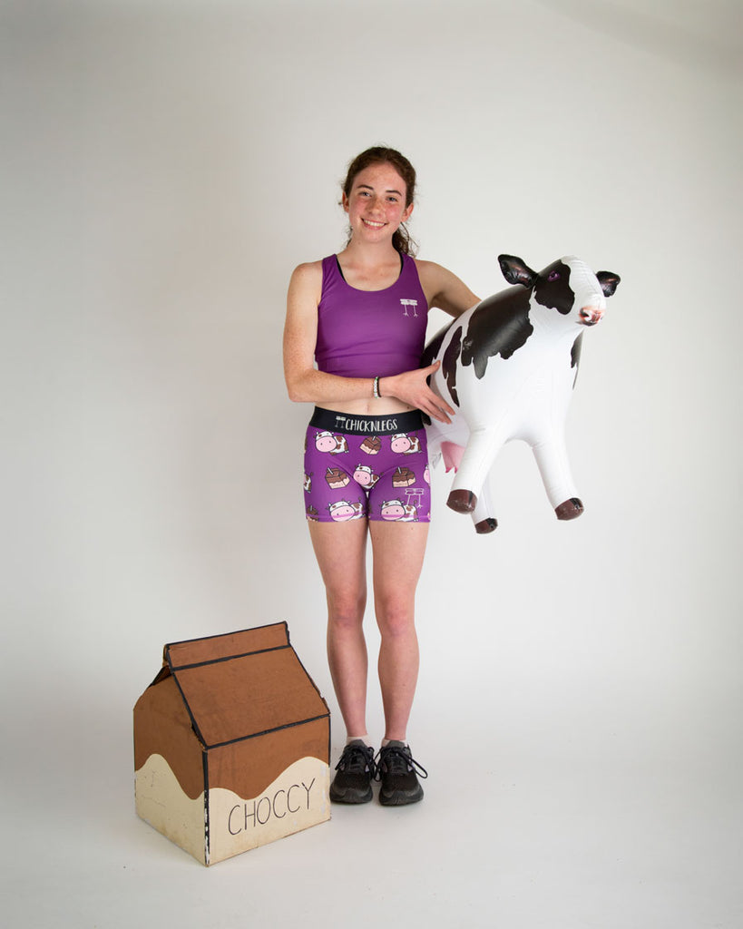 Runner is wearing ChicknLegs in the women's 3 in compression spandex shorts in the choccy cows print. The pattern has chocolate brown cows and chocolate milk on the shorts with a purple background. The model is holding a cow with a huge milk carton next to them.